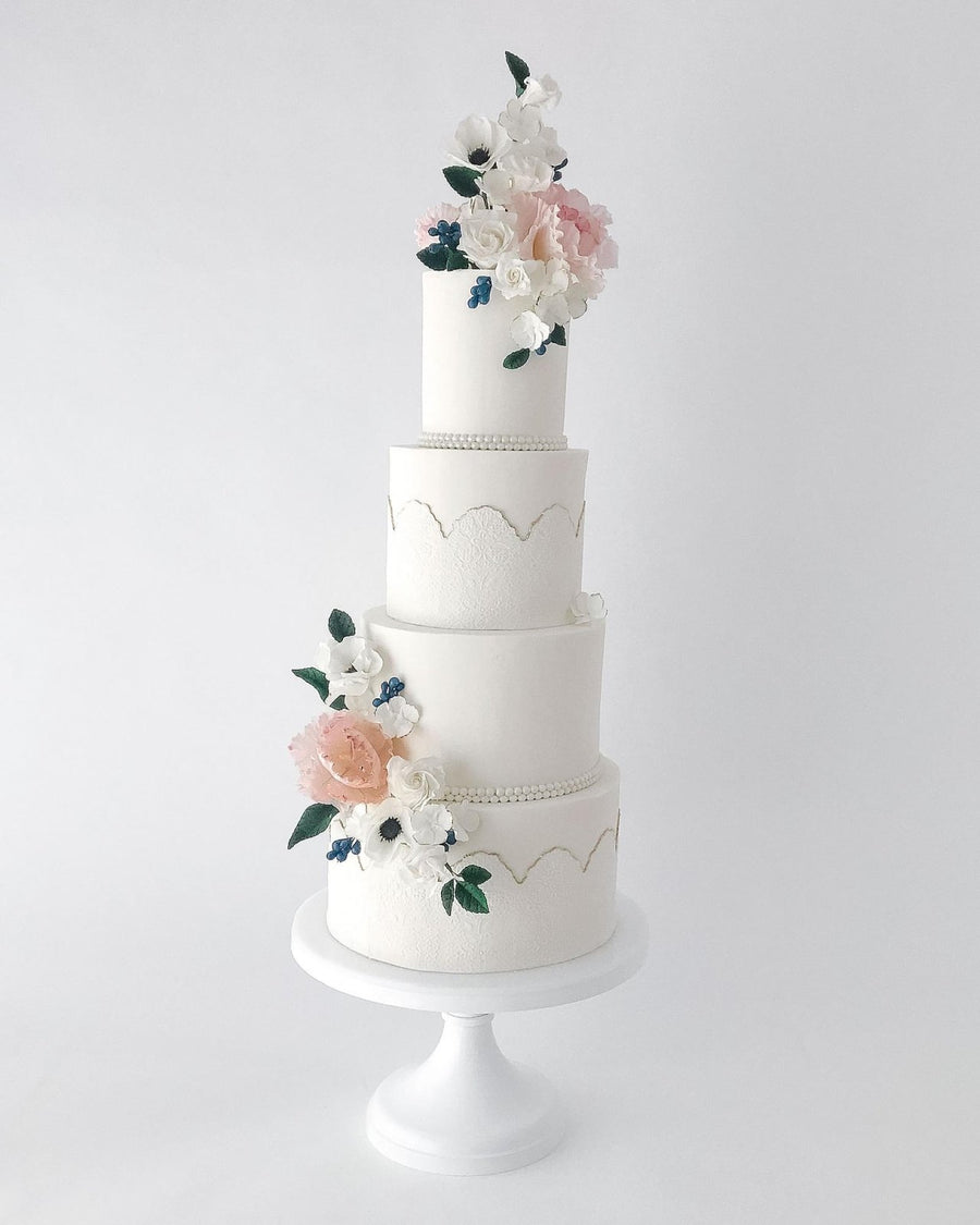 A four-tiered white wedding cake decorated with delicate pastel flowers and greenery, featuring subtle gold accents and pearl-like borders, displayed on a white stand against a plain background.