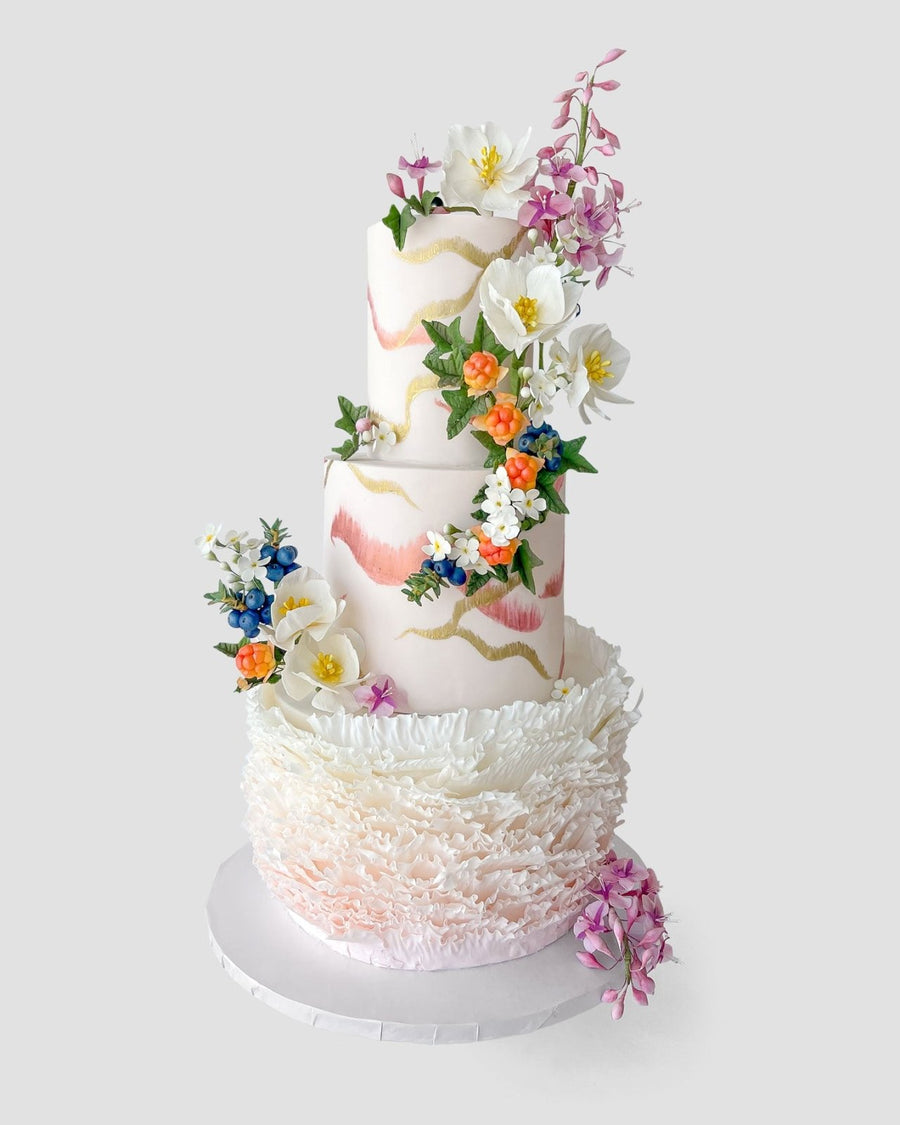 A three-tiered white cake decorated with pink and gold brush strokes, adorned with colorful, realistic sugar flowers and intricate white ruffles around the base. The cake sits on a plain white round board.