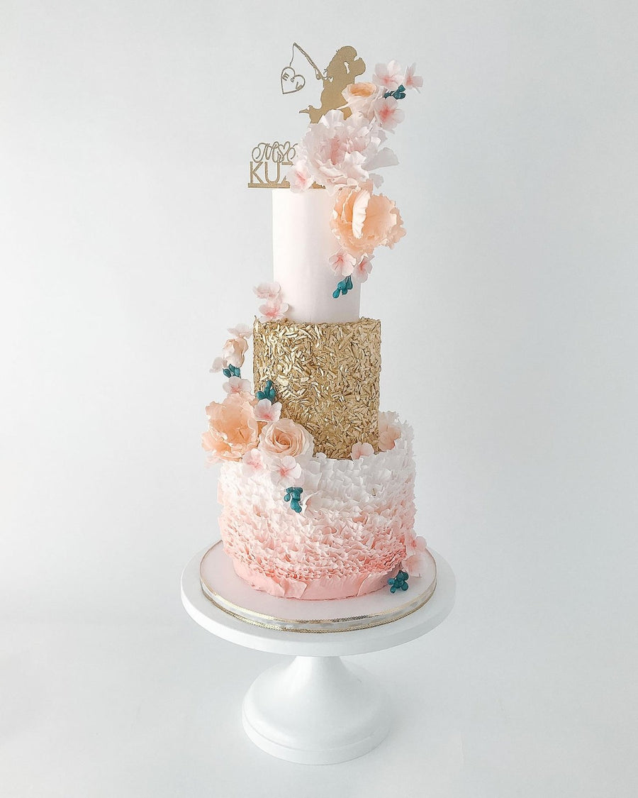 A three-tiered cake with pink, gold, and white layers, decorated with ruffled icing, pastel flowers, a gold cupid topper, and blue butterfly decorations, displayed on a white cake stand against a white background.