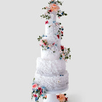 A five-tier, tall white Wedding Cakes $1,000+ by Jenna Rae Cakes features pastel sugar flowers, green leaves, and textured icing, beautifully displayed against a plain light gray background.
