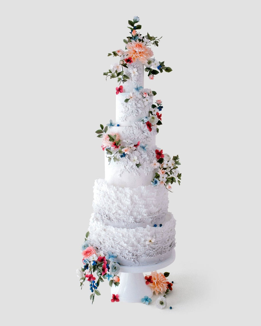 A five-tier, tall white Wedding Cakes $1,000+ by Jenna Rae Cakes features pastel sugar flowers, green leaves, and textured icing, beautifully displayed against a plain light gray background.