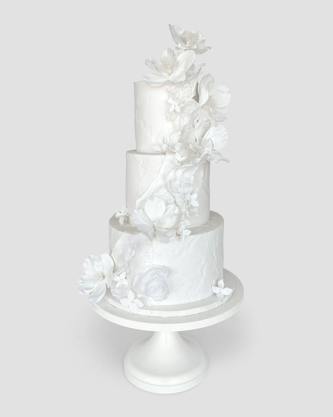A three-tiered white cake decorated with delicate, white sugar flowers cascading down the side, displayed on a simple white cake stand against a plain light gray background.