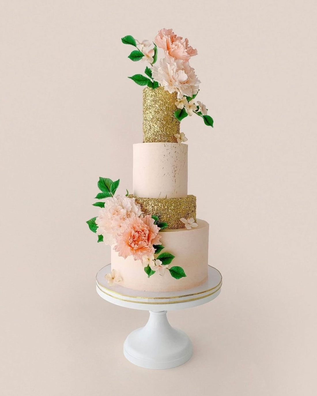 A three-tiered cake with blush pink and glittery gold layers, decorated with large pink flowers and green leaves, displayed on a white cake stand against a light background.