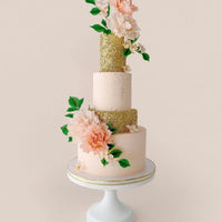 A three-tiered cake with blush pink and glittery gold layers, decorated with large pink flowers and green leaves, displayed on a white cake stand against a light background.